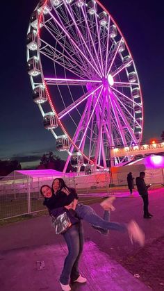 two people posing for a photo in front of a ferris wheel at night with the lights on