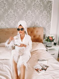 a woman sitting on top of a bed wearing sunglasses and holding a cup in her hand