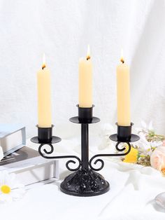 three candles are lit on a black candle holder with flowers in the background and a white sheet behind it