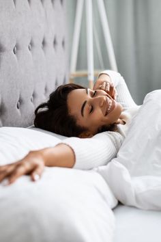 a woman laying on top of a white bed next to a gray headboard and pillows