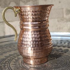 a copper colored pitcher sitting on top of a glass table with a snake in it's mouth