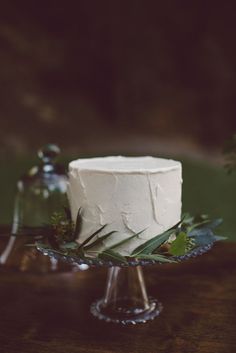 a white frosted cake sitting on top of a wooden table next to greenery