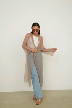 a woman standing in front of a white wall wearing a long cardigan and jeans