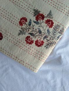 an embroidered piece of cloth with red flowers and leaves on white fabric, sitting on a bed