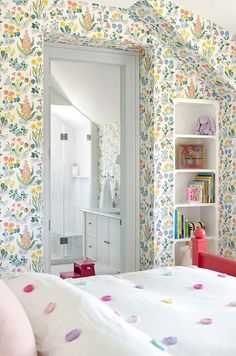 a bedroom with colorful wallpaper and white bed in the foreground is a bookcase full of children's books