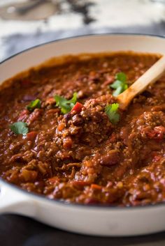 a white bowl filled with chili and meat on top of a table next to a wooden spoon