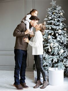 a man and woman kissing in front of a christmas tree with a small child on their shoulders