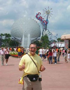 a man standing in front of a fountain at a theme park with his thumbs up