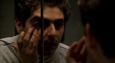 a man looking in the mirror with his hand on his face and another person's reflection behind him