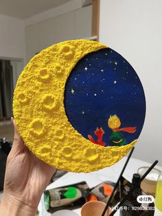 a hand holding up a yellow painted moon with a little boy sitting on the moon