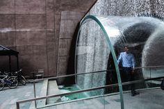 a man standing in front of a water fountain next to bicycles and a building with a waterfall