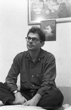 a black and white photo of a man in glasses sitting on a bed with his legs crossed