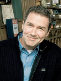 a man in a blue shirt and black jacket smiles at the camera while standing next to shelves with books