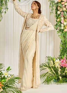 Featuring an ivory-hued shimmer georgette pre-draped saree paired with a U-neckline bustier detailed with pearl, bead, and sequin work. The look is finished with an embellished long cape featuring pearl tassels and a belt. This ensemble is a ready to wear saree for your ease ! Composition : Saree: Georgette, Blouse: Silk, Cape & Belt: Crepe Care: Dry Clean Only and Vacuum Storage All products can be customised for sleeves, length of blouse and neck design Delivery : 4-6 weeks as the product is h Cinderella Fashion, Saree Jacket, Maggam Designs, Embroidery Geometric, Jacket Designs, Embroidered Cape, Fancy Sarees Party Wear, Dress Book, Padded Blouse