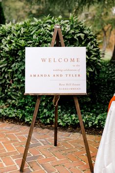 a welcome sign is on an easel in front of some bushes and trees at a wedding