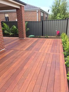 a wooden deck in front of a brick house with plants and shrubs on the side