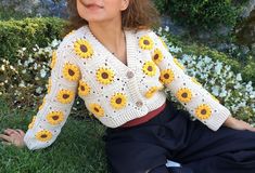 a woman sitting on the grass wearing a sweater with crochet sunflowers