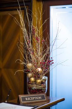 a wooden table topped with a vase filled with red and gold ornaments next to a sign