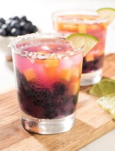 two glasses filled with fruit and ice on top of a wooden cutting board next to limes