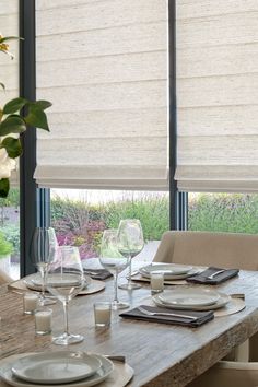 a dining room table with place settings and wine glasses in front of the window that has roman shades on it