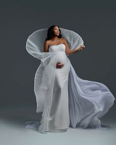 a pregnant woman wearing a white gown and holding an umbrella in her right hand while standing against a gray background