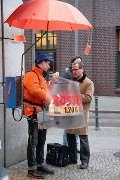 two men are standing under an umbrella on the side of a building while one man is handing something to another man