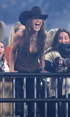 a woman in a cowboy hat is holding a camera and laughing while standing behind a fence