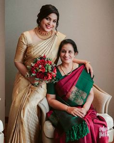 two women in sari sitting next to each other on a white chair and one is holding a bouquet of flowers