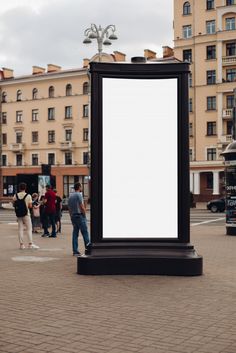 people are walking by a large sign in the middle of a city square with tall buildings behind it