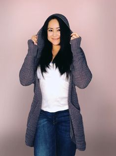 a woman with long black hair wearing a gray cardigan sweater and jeans, standing in front of a pink background