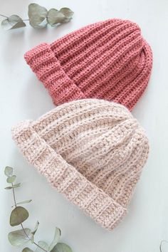 two knitted hats laying next to each other on top of a white table with eucalyptus leaves