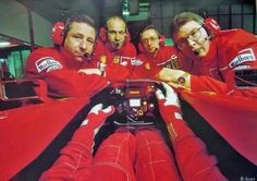 four men in red racing suits are sitting on a car and one man is holding the steering wheel