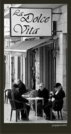 three men sitting at a table in front of a building with a sign that says la dolce vita