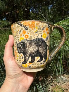 a hand holding a coffee cup with a bear painted on the side and flowers around it