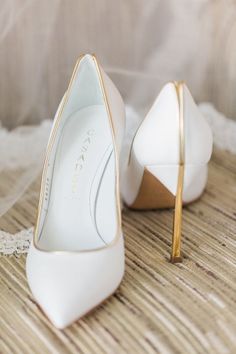 a pair of white high heel shoes with gold detailing on the toes and bottom, sitting on a wooden surface