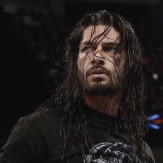 a man with long hair standing in the middle of a wrestling ring wearing a black t - shirt