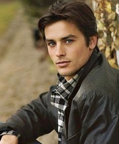 a young man wearing a black jacket and plaid tie sitting on a motorcycle with his hand on the handlebars