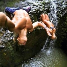 two shirtless men hanging out in the water next to a rock face down waterfall