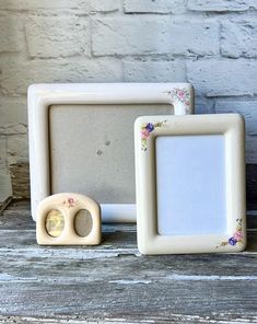 three white frames sitting on top of a wooden table next to a brick wall with flowers painted on them