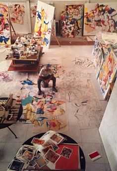 an artist is sitting in his studio surrounded by paintings and other art work on the floor