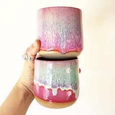 a hand holding two colorful cups in each other's hands, one with pink and blue glaze on it