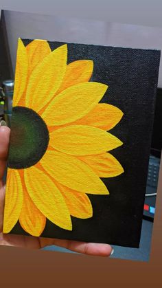 a hand holding up a painted sunflower on a black canvas with yellow and green center