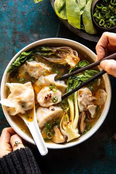 two hands holding chopsticks over a bowl of dumplings with greens and mushrooms