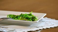 a white bowl filled with green vegetables on top of a lace table cloth next to a fork