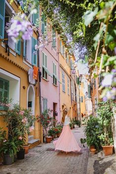 a woman in a pink dress is walking down an alleyway with potted plants