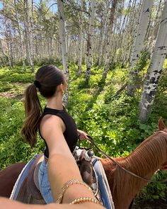 a woman riding on the back of a brown horse through a forest filled with trees