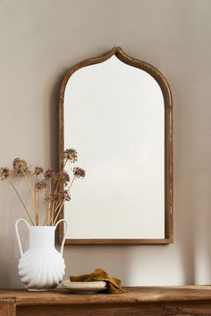 a white vase with dried flowers in front of a mirror on a wooden shelf against a wall