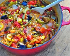 a red pot filled with lots of food on top of a wooden table next to a spoon