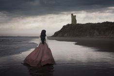 a woman in a long pink dress is standing on the beach looking out at the ocean