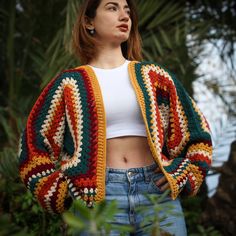 a woman wearing a colorful crochet jacket and jeans standing in front of trees